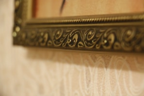A close-up view of an ornate picture frame. The frame features intricate carvings and grooves, with a brass or golden finish. The background consists of a textured wall with a subtle floral pattern in light hues.