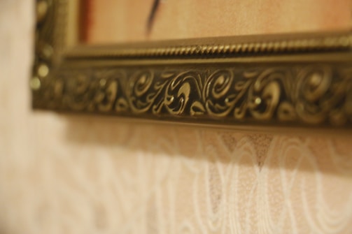 A close-up view of an ornate picture frame. The frame features intricate carvings and grooves, with a brass or golden finish. The background consists of a textured wall with a subtle floral pattern in light hues.