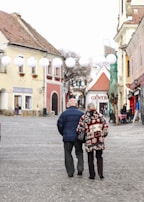A couple walks arm in arm down a cobblestone street lined with charming old buildings. They wear winter clothing, with the woman in a patterned coat and the man in a dark jacket. White lanterns hang above the street, and in the background, ornate architecture and a quaint café ambiance create a cozy atmosphere.