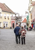 A couple walks arm in arm down a cobblestone street lined with charming old buildings. They wear winter clothing, with the woman in a patterned coat and the man in a dark jacket. White lanterns hang above the street, and in the background, ornate architecture and a quaint caf&eacute; ambiance create a cozy atmosphere.