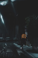 A cyclist rides through a dimly lit city street at night, carrying a bright yellow delivery box. The street is wet, reflecting the light from street lamps, and parked cars line the road. The atmosphere is dark and moody, with limited visibility.