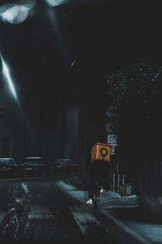 A cyclist rides through a dimly lit city street at night, carrying a bright yellow delivery box. The street is wet, reflecting the light from street lamps, and parked cars line the road. The atmosphere is dark and moody, with limited visibility.