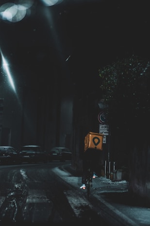 A cyclist rides through a dimly lit city street at night, carrying a bright yellow delivery box. The street is wet, reflecting the light from street lamps, and parked cars line the road. The atmosphere is dark and moody, with limited visibility.