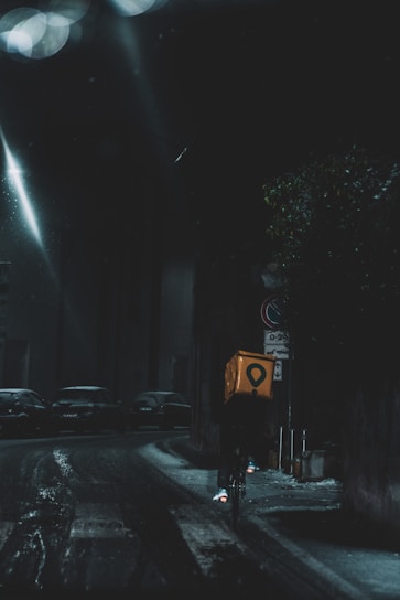 A cyclist rides through a dimly lit city street at night, carrying a bright yellow delivery box. The street is wet, reflecting the light from street lamps, and parked cars line the road. The atmosphere is dark and moody, with limited visibility.