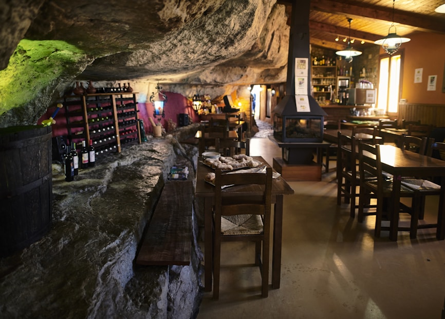 A cozy, rustic interior features a rock ceiling and wooden furniture. Wine bottles are stored on racks along the walls, and a central fireplace adds warmth. Low lighting creates an intimate atmosphere, enhanced by natural stone textures and wooden beams.