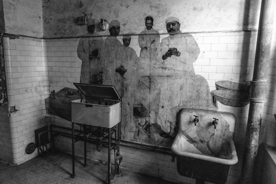 A black and white photograph of an old, worn-out room featuring tiled walls and a vintage sink. The walls display a faded mural of four men in what appear to be medical attire, possibly surgeons from the past. The room has an industrial and antiquated atmosphere with visible pipes and an open metal container on a stand.