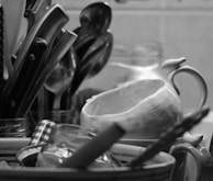 A black-and-white image featuring a close-up of kitchen utensils including knives, spoons, and a ceramic pitcher. Glass jars with checkered lids and other kitchen items are also visible, arranged in a cluttered manner.