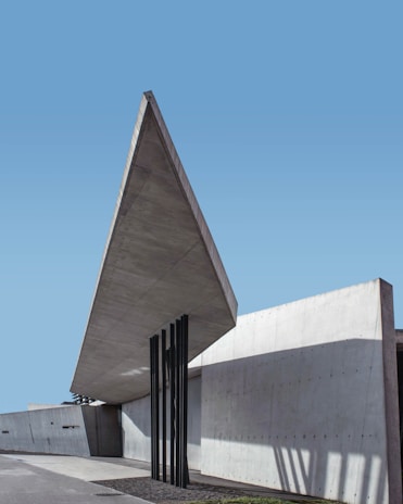 A modern architectural structure with clean, angular lines and a large, cantilevered concrete roof extending outward. Five slender black columns support the overhang. The building's walls are smooth and light gray, contrasting with the bright blue sky. Shadows from the columns add depth and geometric patterns to the scene.