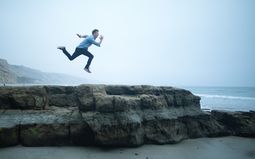 Homem na jaqueta azul pulando na rocha marrom durante o dia