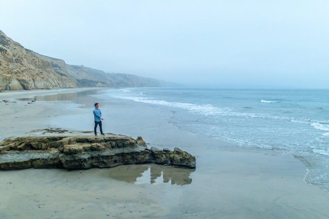 man in blue shirt standing on rock formation by the beach