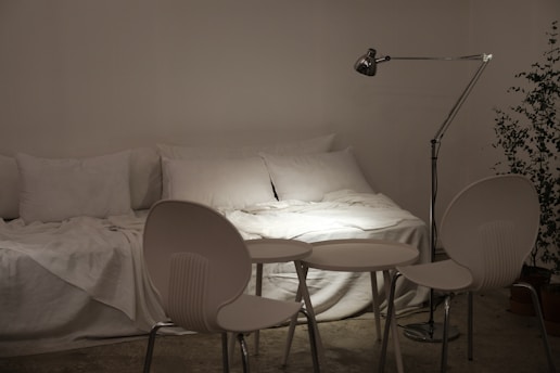 A minimalistic room with a white bed covered in multiple pillows and a soft blanket. In front of the bed are three white chairs surrounding a set of small round tables. To the right, a tall adjustable lamp provides focused light on the bed, and a potted plant adds greenery to the space.