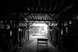 A dimly lit industrial warehouse or workshop with a wooden table in the center and rows of stacked wood along the sides. The ceiling is comprised of exposed beams, and there is minimal light entering through an opening in the background, highlighting some details in the otherwise shadowy interior.
