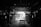 A dimly lit industrial warehouse or workshop with a wooden table in the center and rows of stacked wood along the sides. The ceiling is comprised of exposed beams, and there is minimal light entering through an opening in the background, highlighting some details in the otherwise shadowy interior.