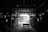 A dimly lit industrial warehouse or workshop with a wooden table in the center and rows of stacked wood along the sides. The ceiling is comprised of exposed beams, and there is minimal light entering through an opening in the background, highlighting some details in the otherwise shadowy interior.