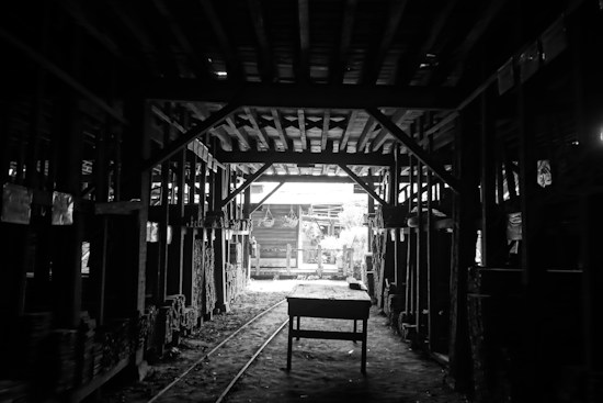 A dimly lit industrial warehouse or workshop with a wooden table in the center and rows of stacked wood along the sides. The ceiling is comprised of exposed beams, and there is minimal light entering through an opening in the background, highlighting some details in the otherwise shadowy interior.