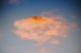 A cloud with soft, fluffy edges is illuminated by a warm, orange glow against a clear blue sky. The light creates a peaceful and serene scene.