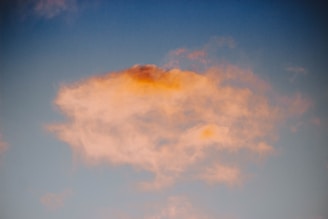 A cloud with soft, fluffy edges is illuminated by a warm, orange glow against a clear blue sky. The light creates a peaceful and serene scene.
