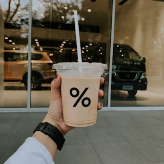 A hand is holding a plastic cup with a logo of a percentage sign. The cup contains a beige-colored beverage and is topped with a clear lid and straw. In the background, there are reflections of cars in the glass windows of a building.
