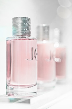 A close-up of three elegant perfume bottles with a focus on the first one in the foreground. The bottles feature a sleek, cylindrical design with a metallic cap and a light pink liquid inside. The background is softly blurred, giving a dreamy, luxurious feel.