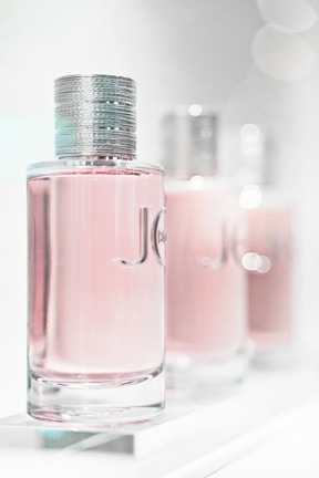 A close-up of three elegant perfume bottles with a focus on the first one in the foreground. The bottles feature a sleek, cylindrical design with a metallic cap and a light pink liquid inside. The background is softly blurred, giving a dreamy, luxurious feel.