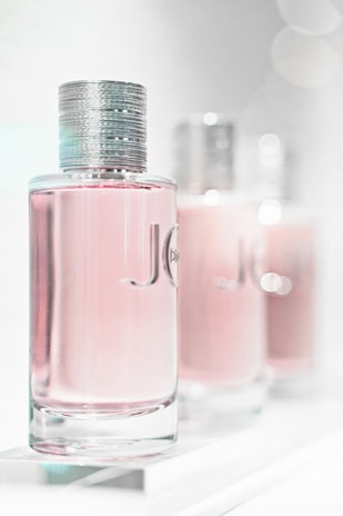 A close-up of three elegant perfume bottles with a focus on the first one in the foreground. The bottles feature a sleek, cylindrical design with a metallic cap and a light pink liquid inside. The background is softly blurred, giving a dreamy, luxurious feel.