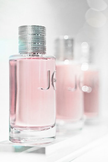 A close-up of three elegant perfume bottles with a focus on the first one in the foreground. The bottles feature a sleek, cylindrical design with a metallic cap and a light pink liquid inside. The background is softly blurred, giving a dreamy, luxurious feel.