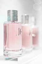 A close-up of three elegant perfume bottles with a focus on the first one in the foreground. The bottles feature a sleek, cylindrical design with a metallic cap and a light pink liquid inside. The background is softly blurred, giving a dreamy, luxurious feel.