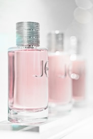 A close-up of three elegant perfume bottles with a focus on the first one in the foreground. The bottles feature a sleek, cylindrical design with a metallic cap and a light pink liquid inside. The background is softly blurred, giving a dreamy, luxurious feel.