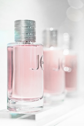 A close-up of three elegant perfume bottles with a focus on the first one in the foreground. The bottles feature a sleek, cylindrical design with a metallic cap and a light pink liquid inside. The background is softly blurred, giving a dreamy, luxurious feel.