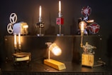A variety of vintage projectors and mechanical objects are creatively transformed into lamps. The setup features six unique lamps, each with its own style and base, including film reels and gears. The lamps emit a warm, ambient light, casting a cozy glow.