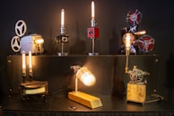 A variety of vintage projectors and mechanical objects are creatively transformed into lamps. The setup features six unique lamps, each with its own style and base, including film reels and gears. The lamps emit a warm, ambient light, casting a cozy glow.