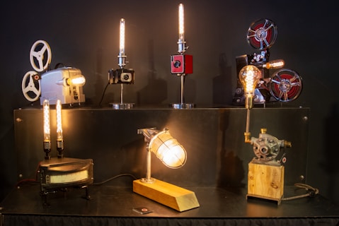 A variety of vintage projectors and mechanical objects are creatively transformed into lamps. The setup features six unique lamps, each with its own style and base, including film reels and gears. The lamps emit a warm, ambient light, casting a cozy glow.