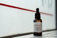 A bottle of beard oil with a dropper cap is placed on a ledge. The label reads 'Josè Manuel Hair Care Beard Oil' and notes the inclusion of mandarin, macadamia, and Abyssinian oil. The background consists of a red stripe on a frosted or translucent surface, allowing a subtle, diffused light to illuminate the bottle.