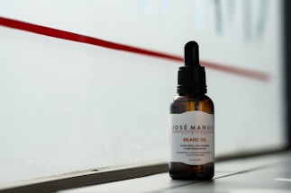 A bottle of beard oil with a dropper cap is placed on a ledge. The label reads 'Josè Manuel Hair Care Beard Oil' and notes the inclusion of mandarin, macadamia, and Abyssinian oil. The background consists of a red stripe on a frosted or translucent surface, allowing a subtle, diffused light to illuminate the bottle.
