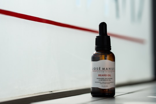 A bottle of beard oil with a dropper cap is placed on a ledge. The label reads 'Josè Manuel Hair Care Beard Oil' and notes the inclusion of mandarin, macadamia, and Abyssinian oil. The background consists of a red stripe on a frosted or translucent surface, allowing a subtle, diffused light to illuminate the bottle.