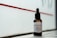 A bottle of beard oil with a dropper cap is placed on a ledge. The label reads 'Josè Manuel Hair Care Beard Oil' and notes the inclusion of mandarin, macadamia, and Abyssinian oil. The background consists of a red stripe on a frosted or translucent surface, allowing a subtle, diffused light to illuminate the bottle.