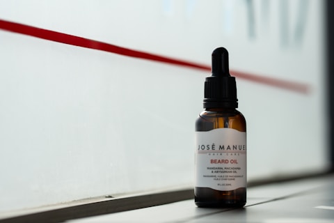 A bottle of beard oil with a dropper cap is placed on a ledge. The label reads 'Josè Manuel Hair Care Beard Oil' and notes the inclusion of mandarin, macadamia, and Abyssinian oil. The background consists of a red stripe on a frosted or translucent surface, allowing a subtle, diffused light to illuminate the bottle.