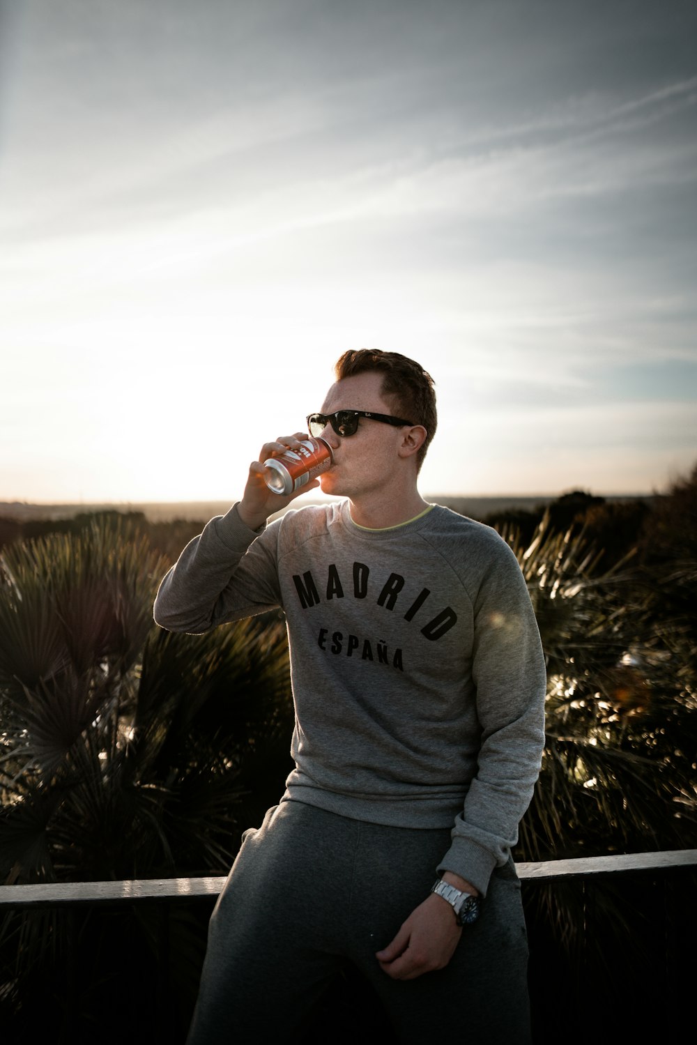man in grey sweater drinking beverage on can while sitting on planter