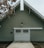 A large barn-style building with a green facade and white trim features a prominent garage door with small windows and decorative paneling. A single outdoor light fixture is situated above the garage door, and the roof has an angled design with scalloped eaves. The ground in front appears to be paved, with brown leaves visible to the side, suggesting an autumn setting.