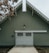 A large barn-style building with a green facade and white trim features a prominent garage door with small windows and decorative paneling. A single outdoor light fixture is situated above the garage door, and the roof has an angled design with scalloped eaves. The ground in front appears to be paved, with brown leaves visible to the side, suggesting an autumn setting.