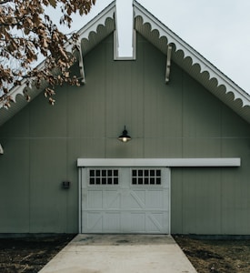A large barn-style building with a green facade and white trim features a prominent garage door with small windows and decorative paneling. A single outdoor light fixture is situated above the garage door, and the roof has an angled design with scalloped eaves. The ground in front appears to be paved, with brown leaves visible to the side, suggesting an autumn setting.