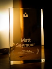 A glass award placed on a wooden surface with engraved text showing the name Matt Seymour and indicating recognition from the Unsplash Community in 2018. The award also features two laurel wreaths and a geometric logo near the top. The background is softly lit, providing a warm ambiance.