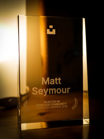 A glass award placed on a wooden surface with engraved text showing the name Matt Seymour and indicating recognition from the Unsplash Community in 2018. The award also features two laurel wreaths and a geometric logo near the top. The background is softly lit, providing a warm ambiance.