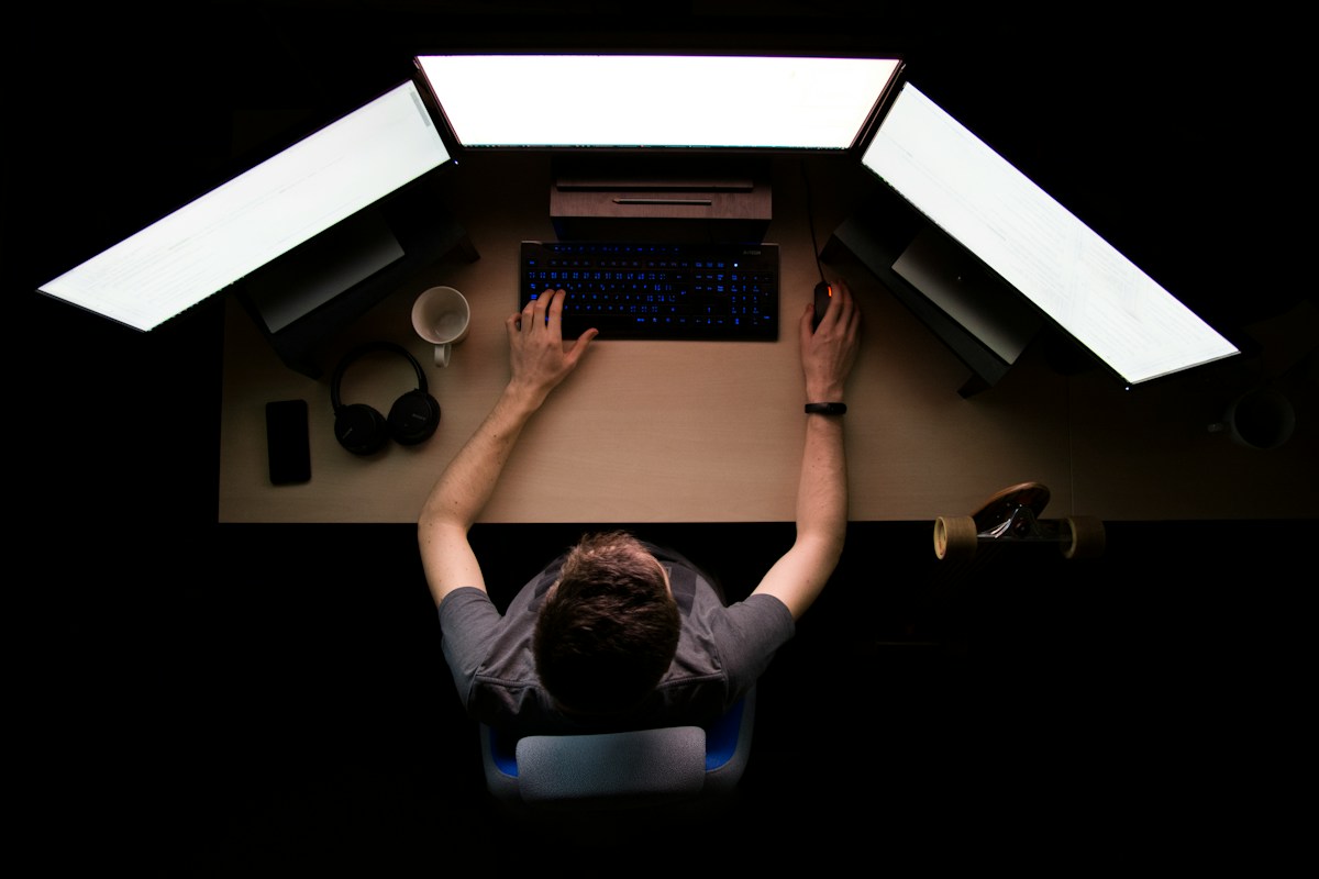 A developer seen from above at a dark desk, hands on a backlit keyboard, three bright monitors glowing around them
