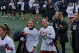 A group of young women, likely a lacrosse team, walk together on a sports field. They are wearing athletic gear, including white shirts and holding lacrosse sticks and water bottles. Some are wearing jackets, and they appear to be in good spirits, smiling and talking with each other.