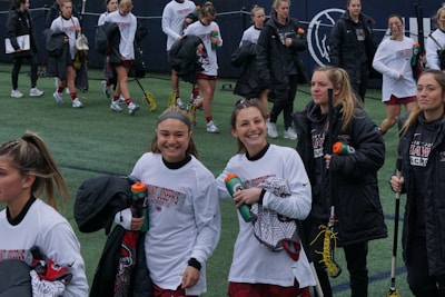 A group of young women, likely a lacrosse team, walk together on a sports field. They are wearing athletic gear, including white shirts and holding lacrosse sticks and water bottles. Some are wearing jackets, and they appear to be in good spirits, smiling and talking with each other.
