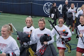 A group of young women, wearing white long-sleeve shirts and maroon shorts, are smiling and walking on a sports field with lacrosse sticks. They appear to be part of a lacrosse team, indicated by the text on their shirts. Some are holding water bottles and jackets. The field is lined with artificial turf and part of a stadium or sports complex is visible in the background.