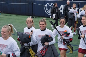 A group of young women, wearing white long-sleeve shirts and maroon shorts, are smiling and walking on a sports field with lacrosse sticks. They appear to be part of a lacrosse team, indicated by the text on their shirts. Some are holding water bottles and jackets. The field is lined with artificial turf and part of a stadium or sports complex is visible in the background.