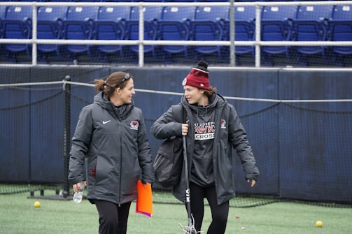Two people are dressed warmly in athletic gear, standing on a field and engaging in conversation. One individual has a clipboard and a water bottle, while the other carries a sports stick. They appear relaxed and friendly.