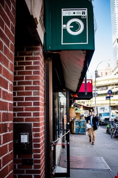 A city street scene with a brick building and a dark green awning featuring a laundry machine illustration. Below the awning, there is a glass door with a pull sign, and a man is walking on the sidewalk holding a coffee cup. The background includes urban elements like a street sign, bicycles, and parked vehicles.